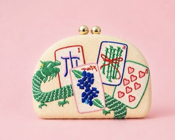 A small cream-colored clutch purse with a gold-tone clasp, decorated with embroidered playing cards, a green dragon, and blue flowers, set against a solid pink background.